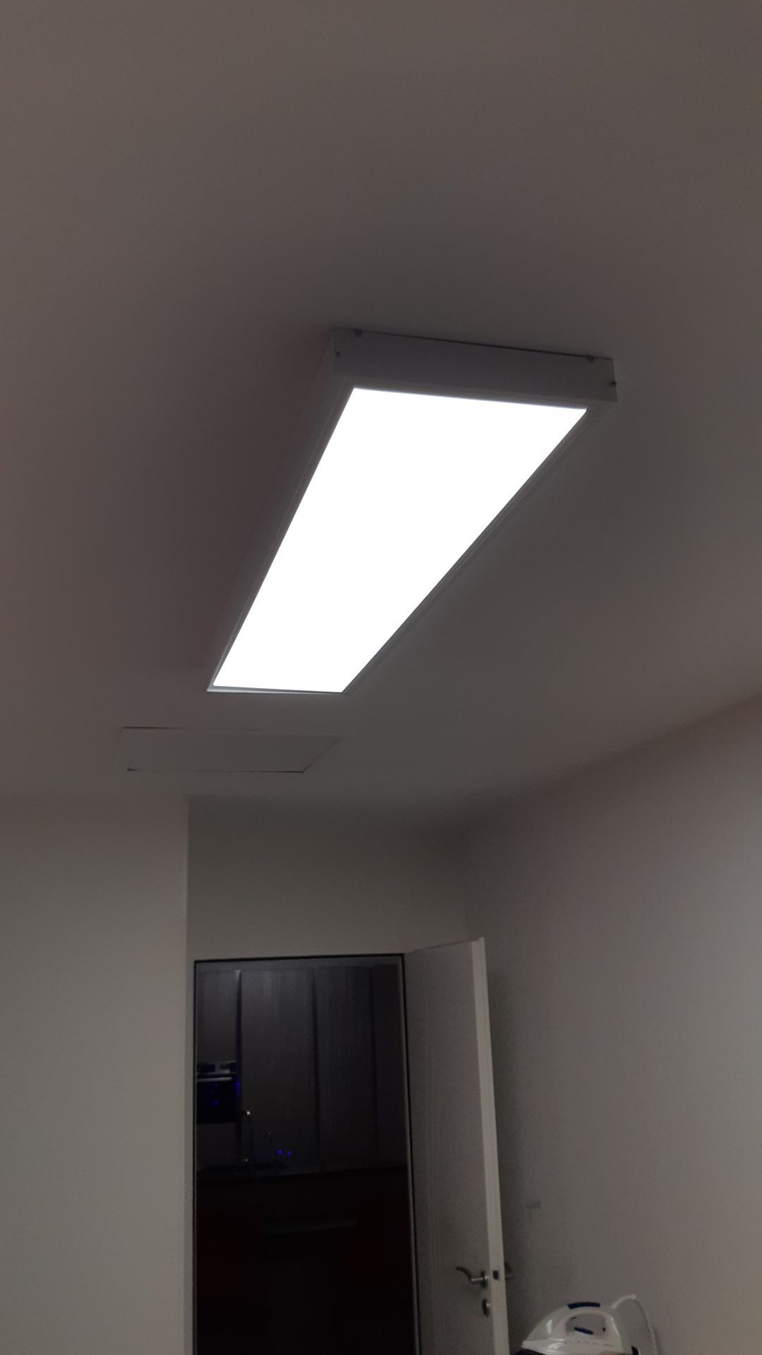 Plafonnier rectangulaire et lumineux dans un couloir avec une porte sombre et des murs blancs.