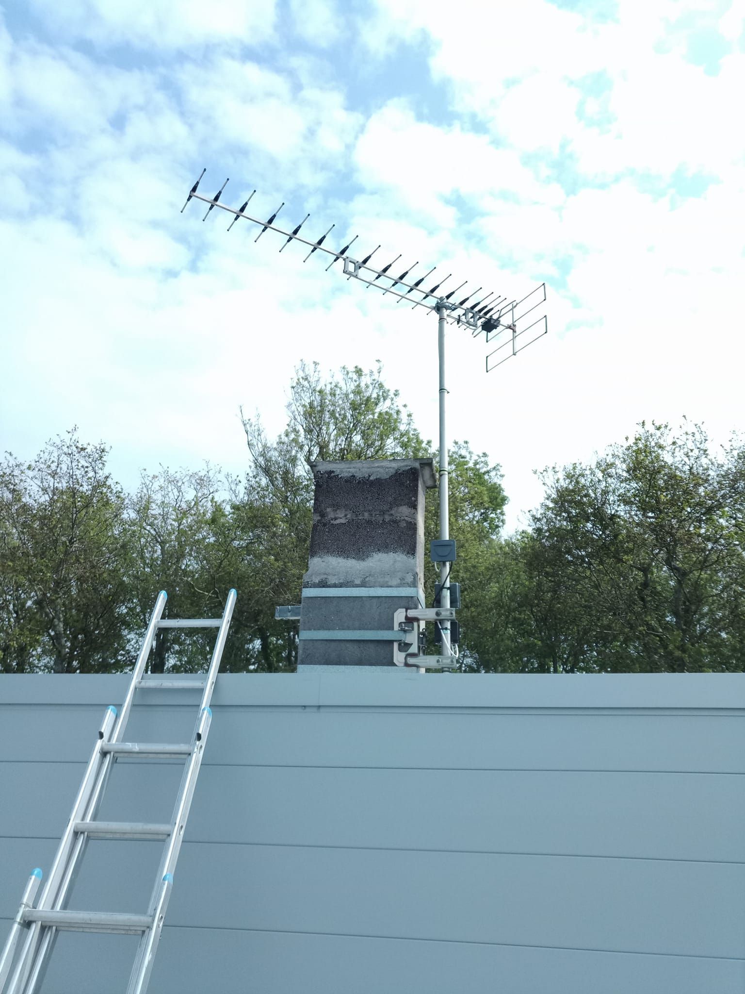 Échelle contre un toit bleu avec une cheminée tenant une antenne de télévision contre un ciel nuageux.