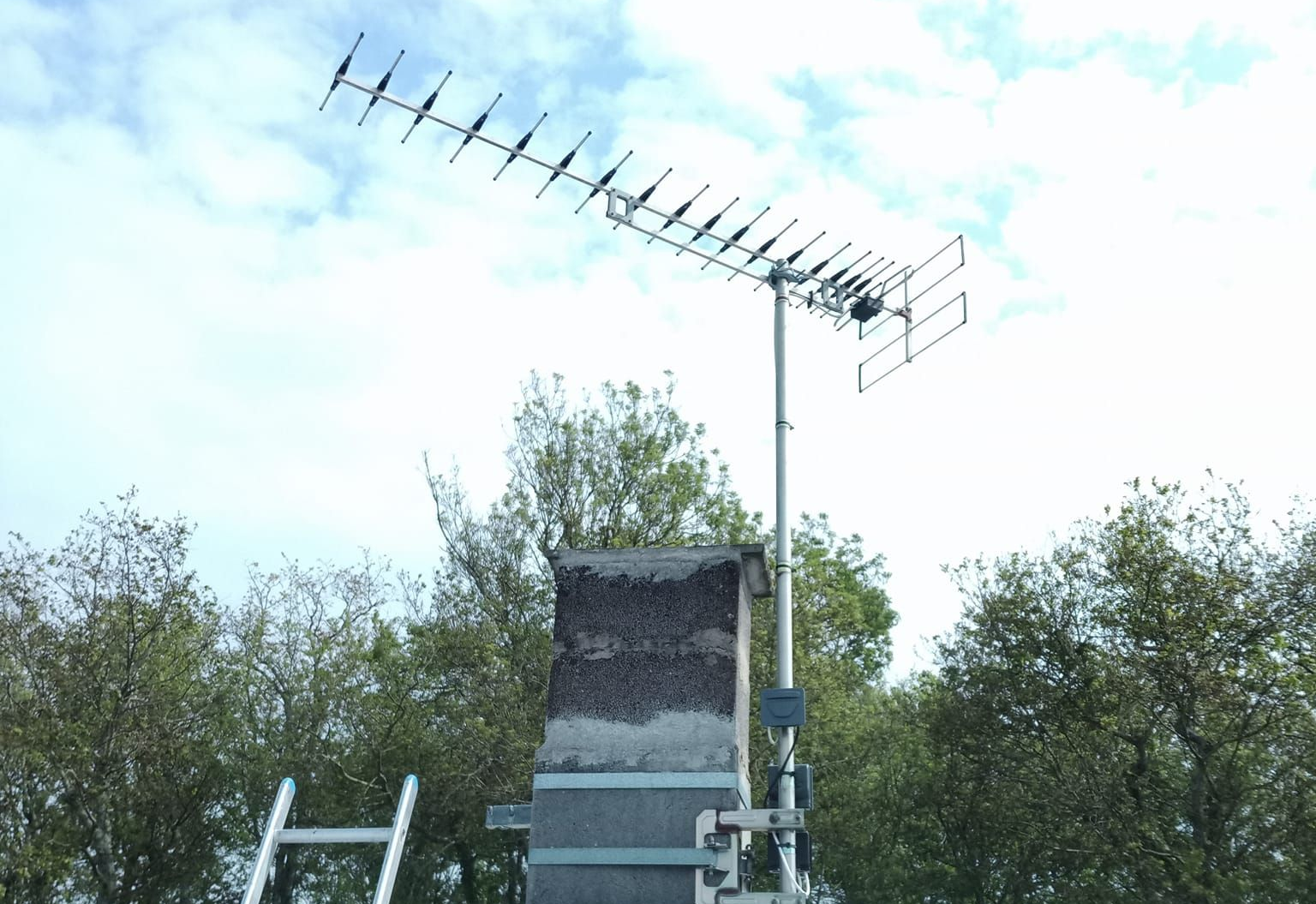 Antenne de télévision montée sur une cheminée, avec des arbres et le ciel en arrière-plan.