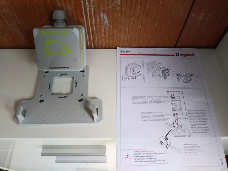 Boîte électrique grise, support de montage et instructions sur une étagère blanche.