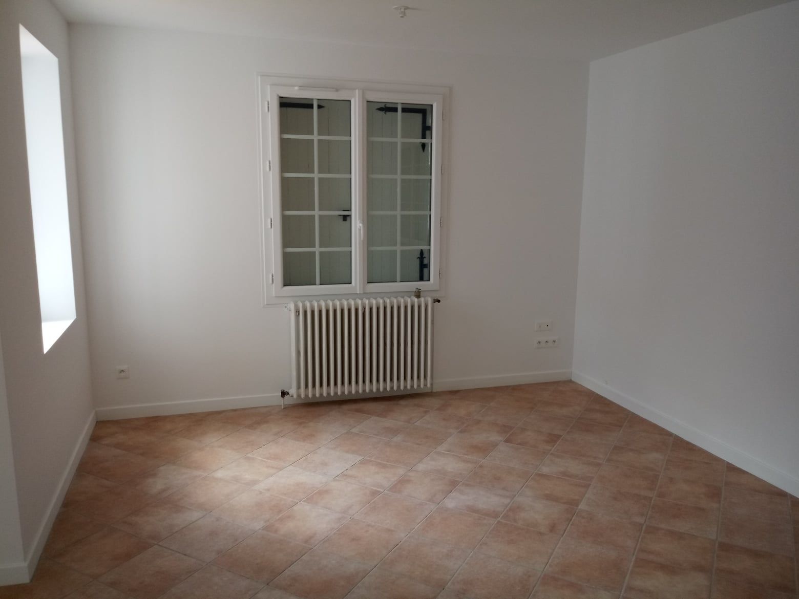 Pièce vide avec fenêtre, radiateur et carrelage au sol. Les murs sont blancs.