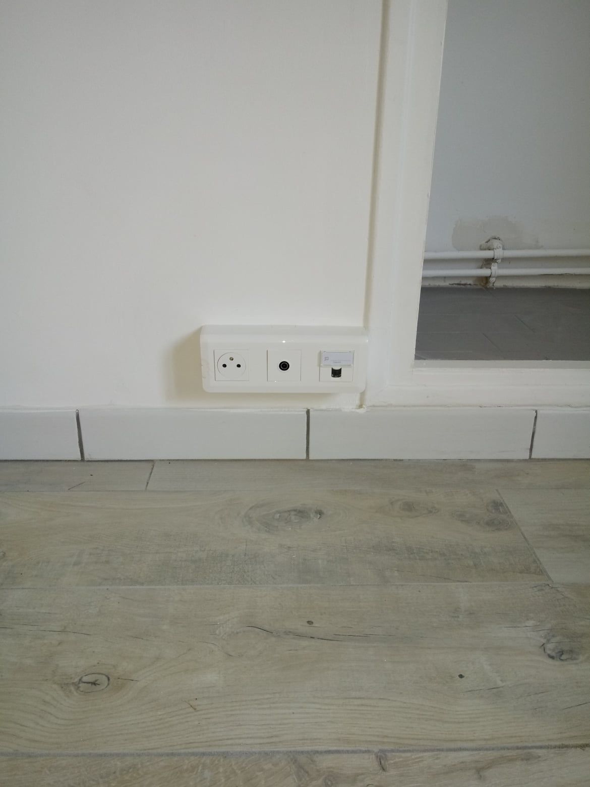 Mur blanc avec une prise de courant, une prise téléphonique et un port de données au-dessus d'un plancher en bois clair.