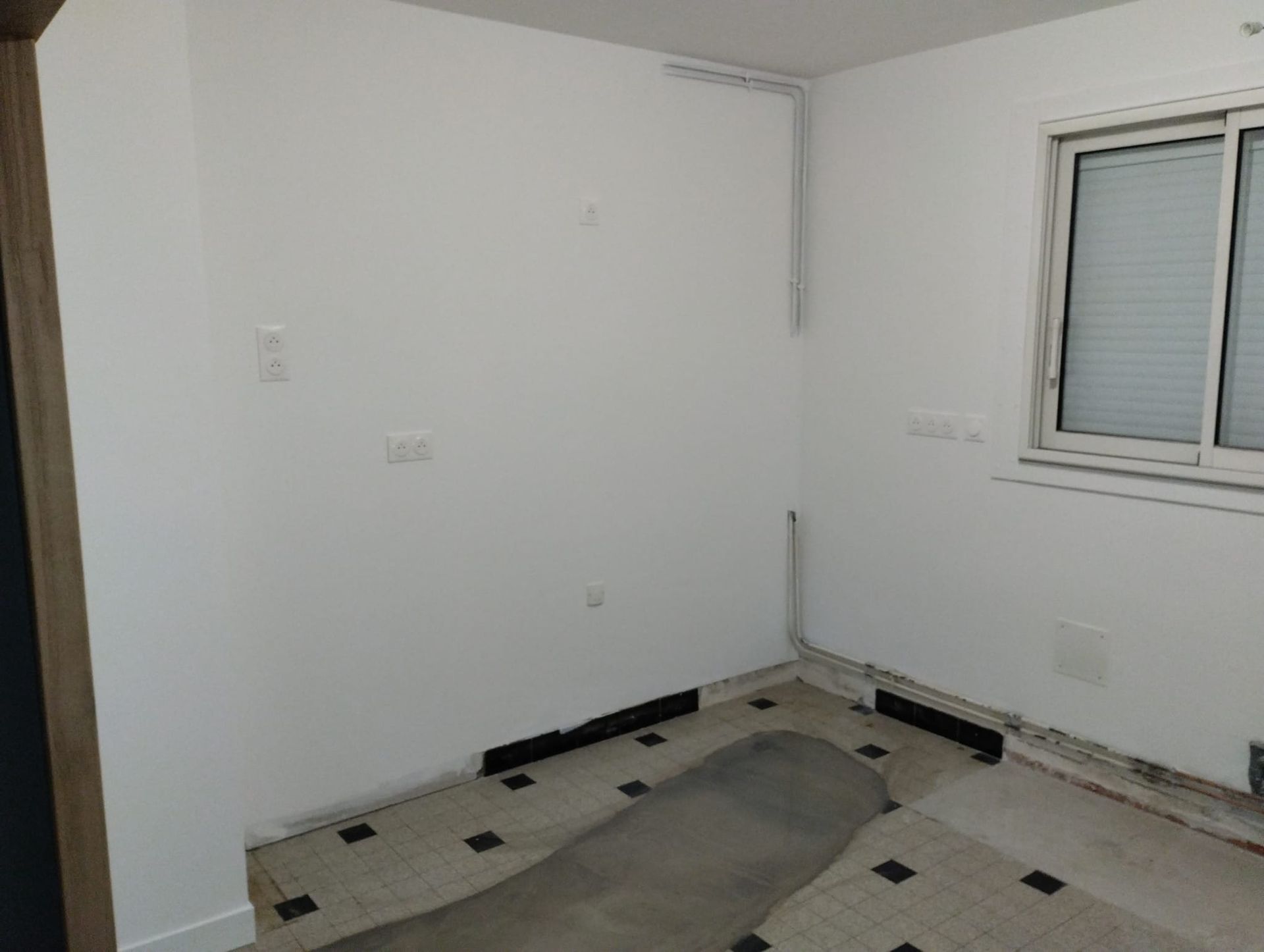 Pièce vide avec murs blancs, fenêtre avec stores et sol en damier.