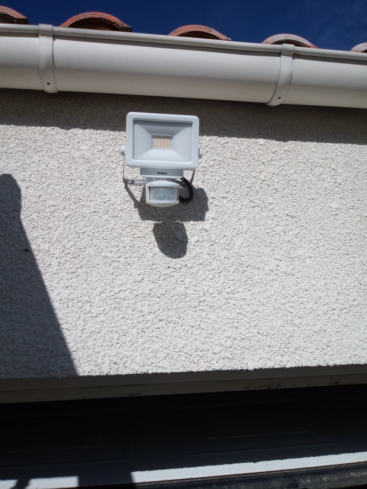 Projecteur LED blanc à détection de mouvement monté sur un mur extérieur en stuc sous une gouttière.