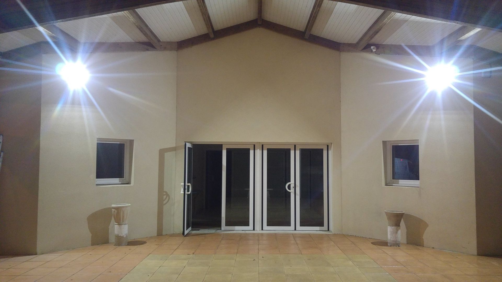 Extérieur d'un bâtiment la nuit. Murs beiges, grande porte vitrée et deux lumières vives.
