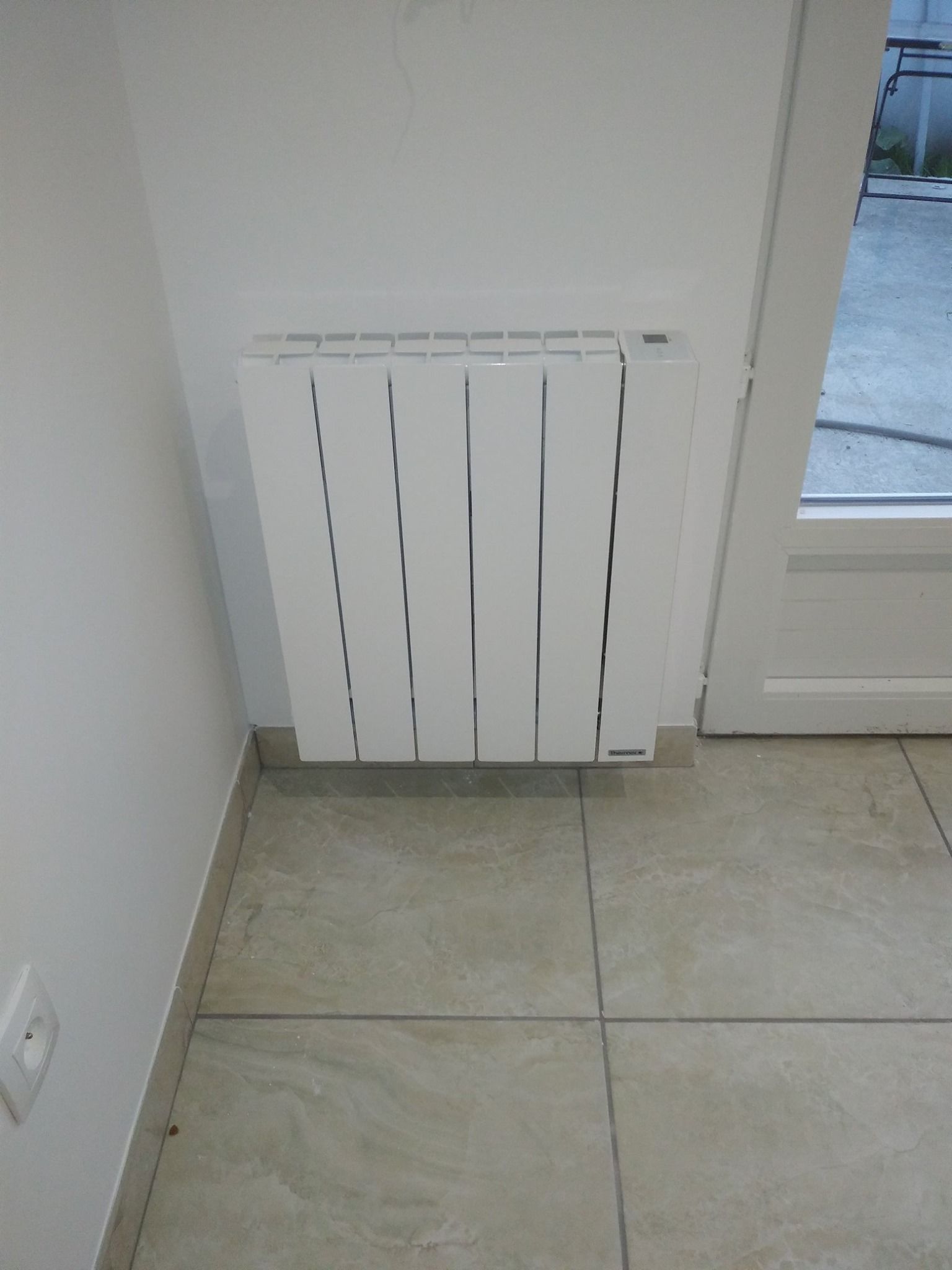 Radiateur électrique blanc sur un mur blanc dans une pièce avec sol carrelé.