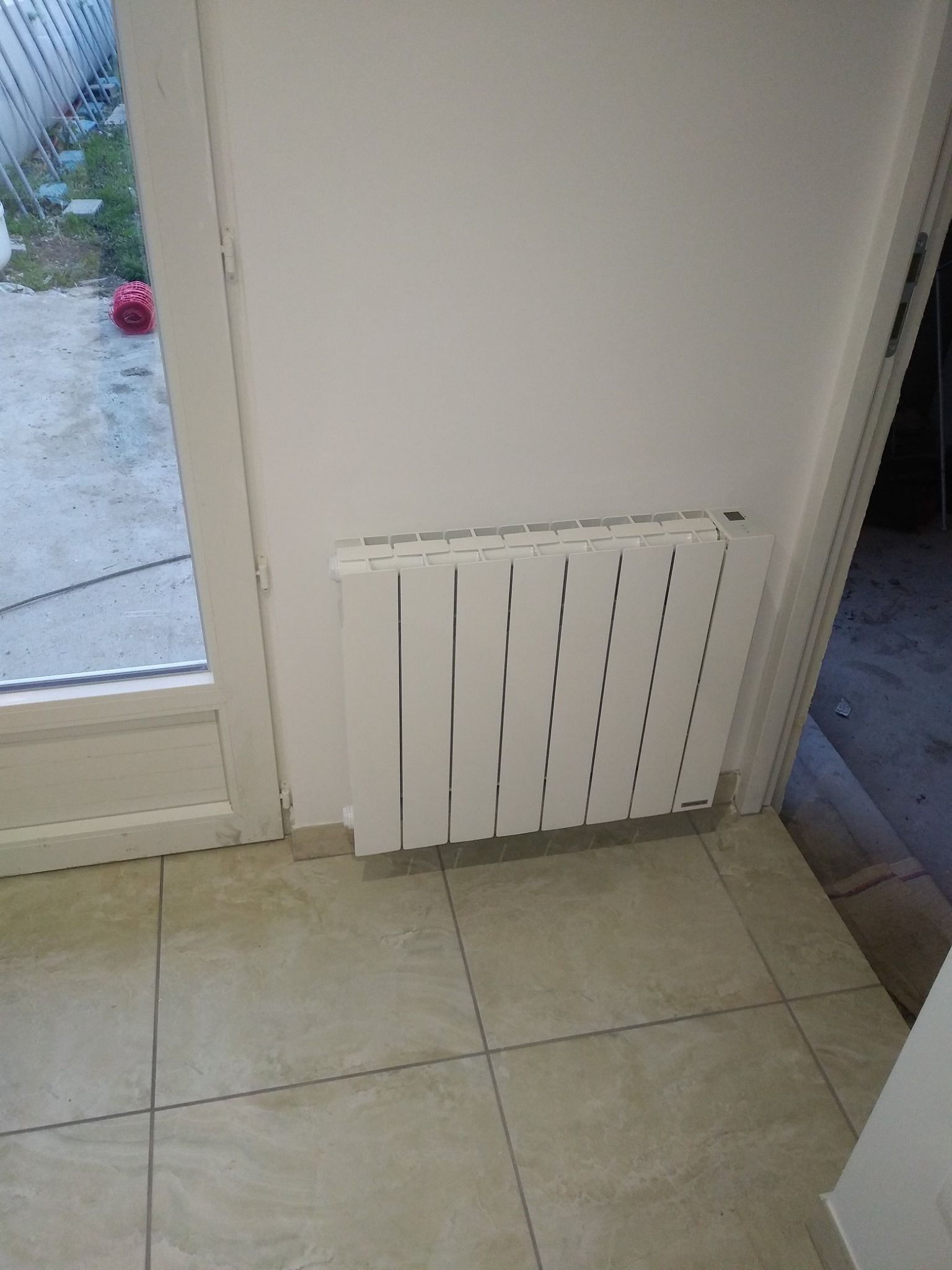 Radiateur blanc fixé sur un mur blanc, à côté d'une porte vitrée. Carrelage beige au sol.