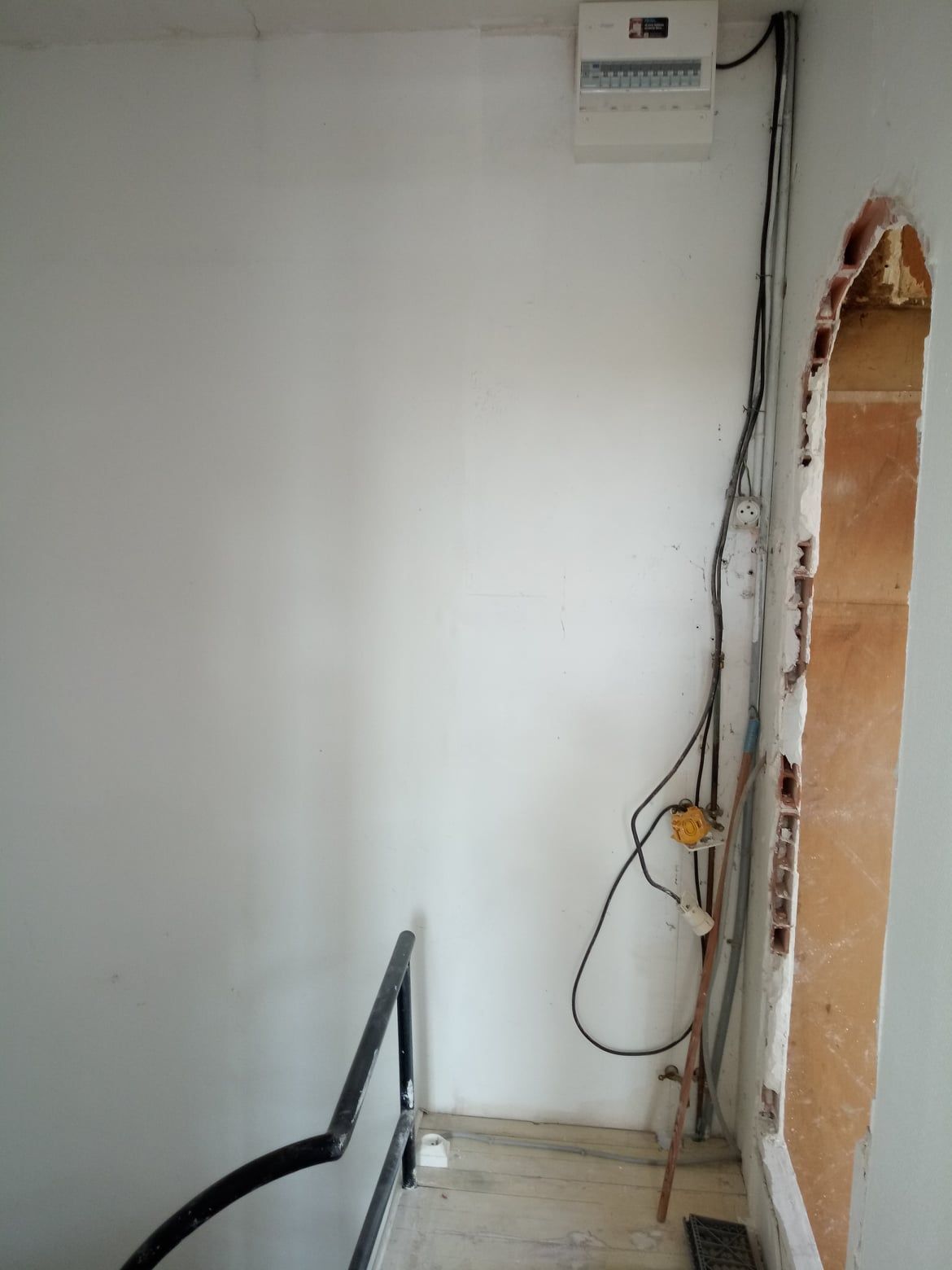 Mur blanc avec fils apparents, tableau électrique et porte. Rampe d'escalier visible en bas à gauche.