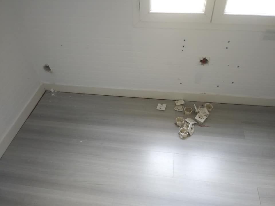 Intérieur de la pièce avec des trous dans le mur blanc, un sol gris et des débris près de la fenêtre.