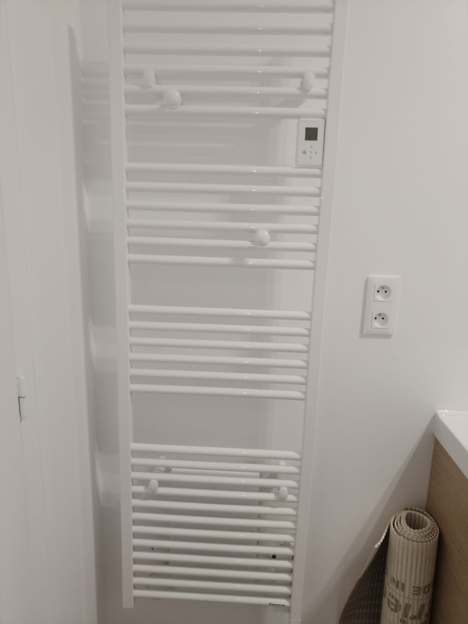 Chauffe-serviettes blanc sur un mur blanc, avec un panneau de commande numérique et une prise électrique à proximité.