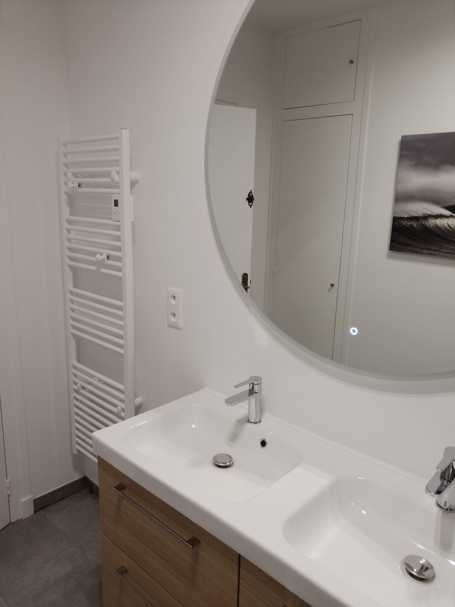 Salle de bains avec lavabo blanc, meuble en bois, grand miroir rond et radiateur sèche-serviettes.
