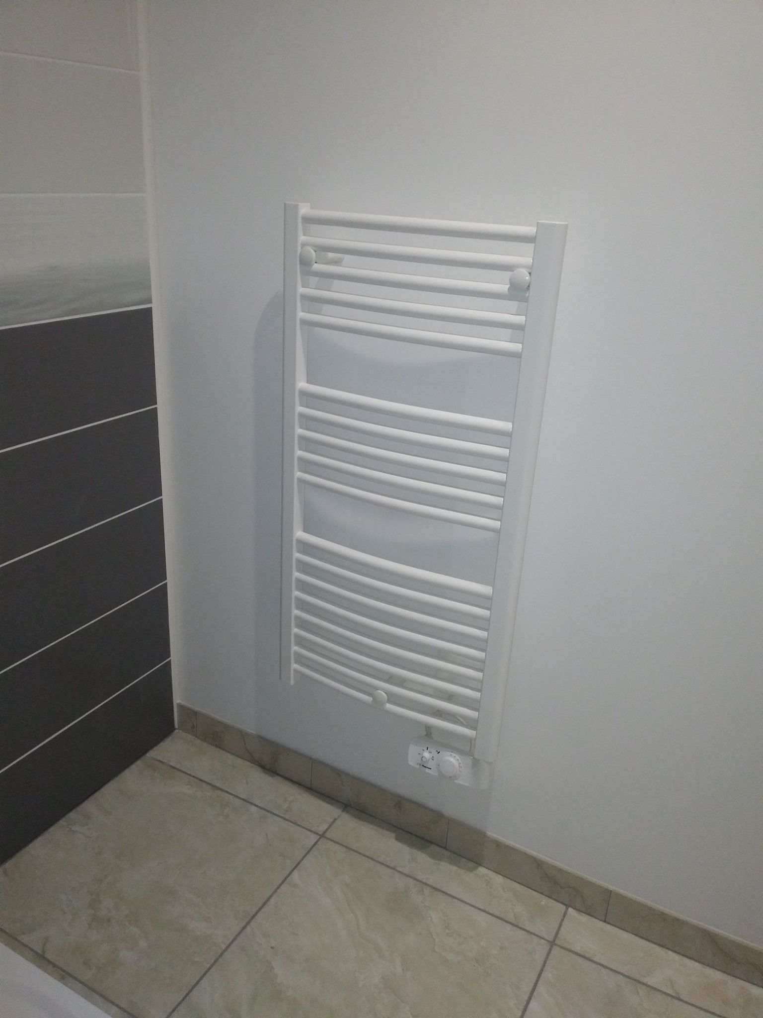 Chauffe-serviettes blanc sur un mur gris clair dans une salle de bains avec sol carrelé.