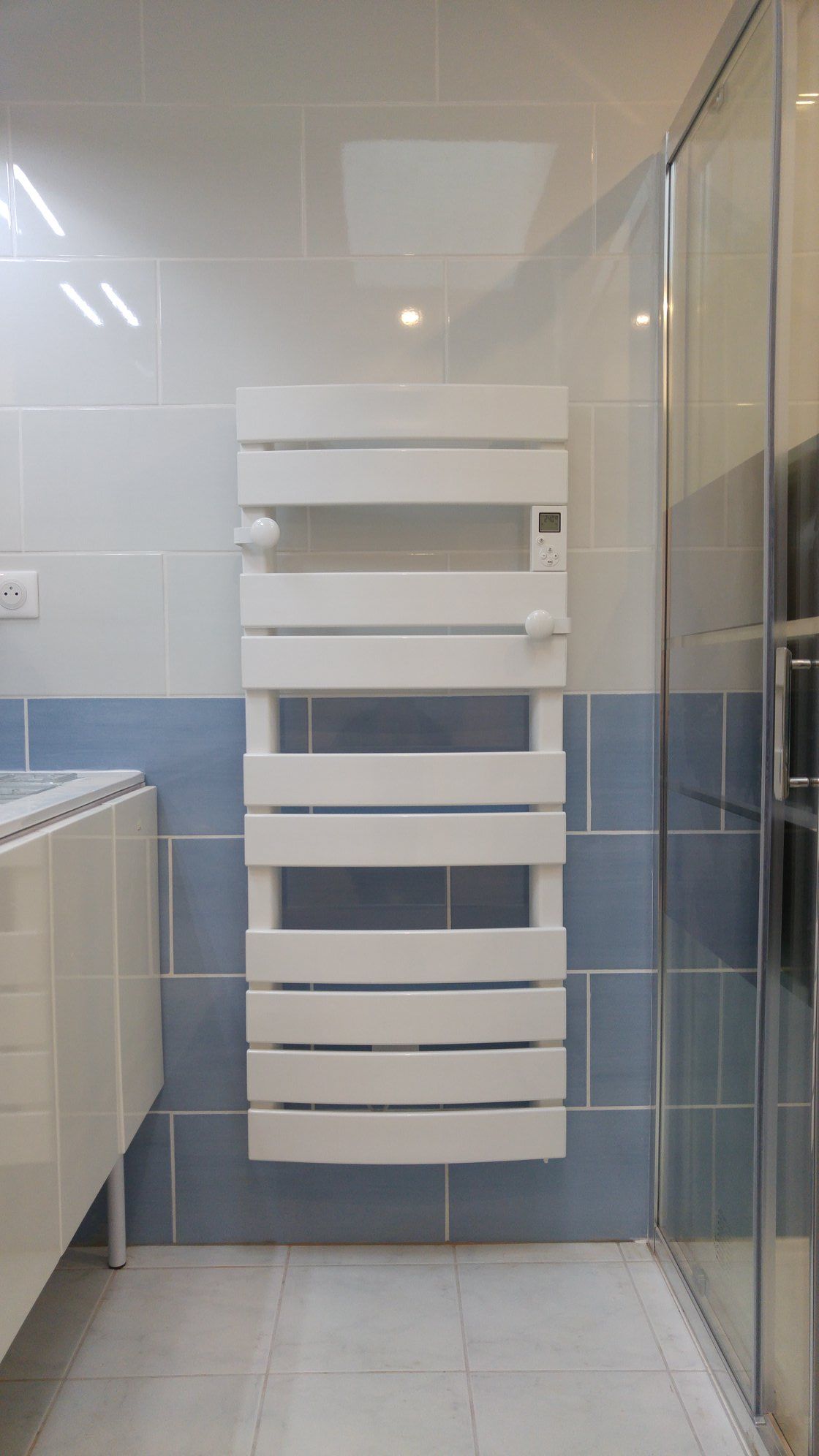 Chauffe-serviettes blanc sur un mur carrelé bleu dans une salle de bains, à côté d'une douche.