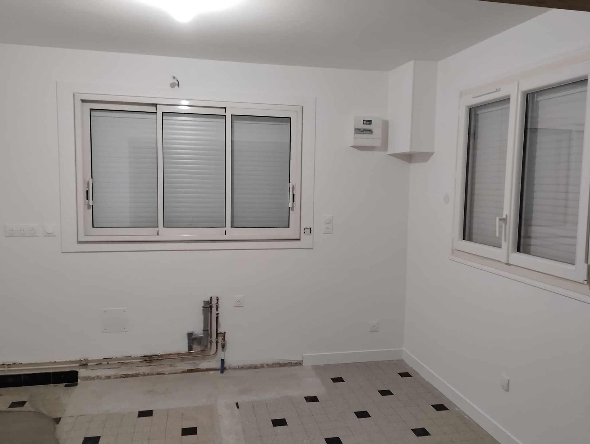 Pièce blanche vide avec deux fenêtres, un local technique et un sol carrelé.