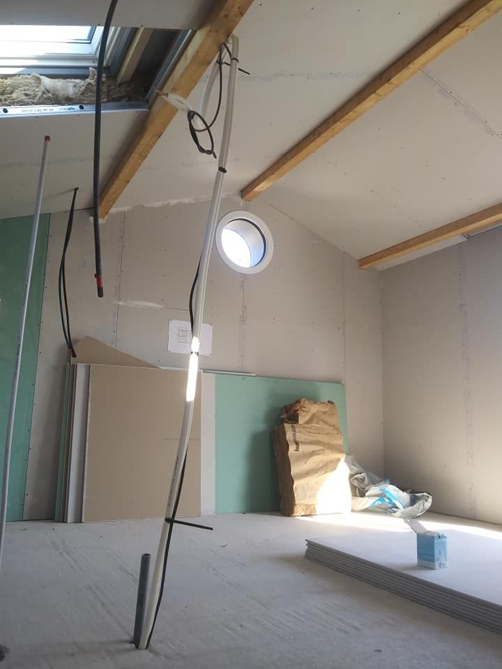 Intérieur d'une pièce inachevée avec cloisons sèches, poutres apparentes et fenêtre ronde. Câbles et matériaux présents.