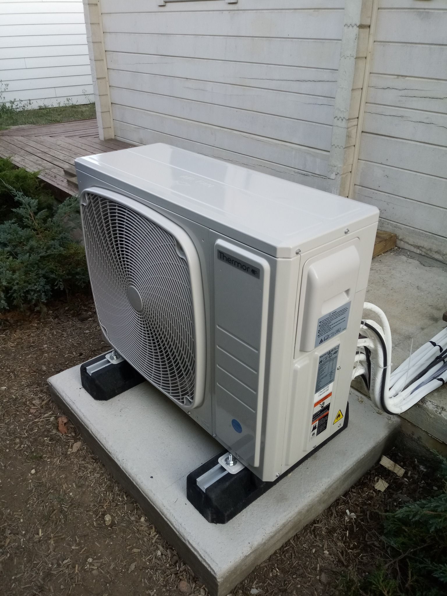 Unité de pompe à chaleur extérieure sur une dalle de béton, contre une clôture en bois blanche.