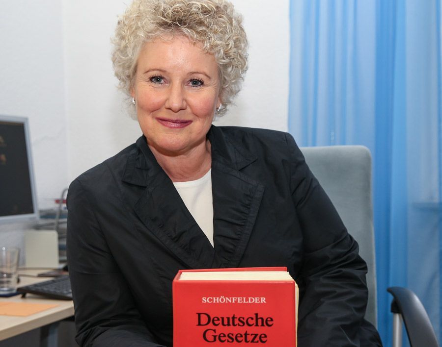 Eine Frau im Anzug sitzt an einem Schreibtisch und schreibt auf ein Stück Papier