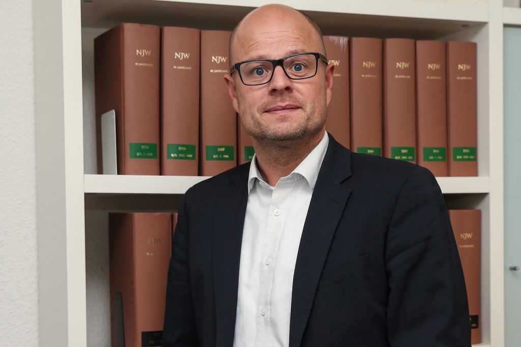 Ein Mann im Anzug und mit Brille steht vor einem Bücherregal voller Bücher.