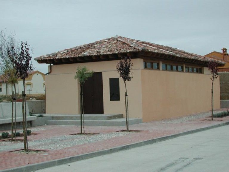 Edificio de color canela, con techo de tejas, puerta oscura y ventanas pequeñas, con árboles jóvenes a lo largo de la acera de ladrillo.