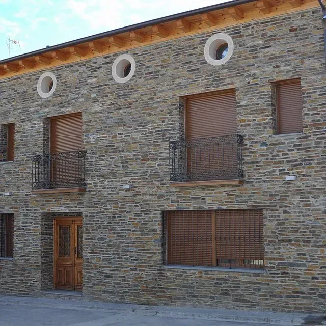 Edificio con fachada de piedra, con puerta de madera, contraventanas y pequeños balcones de hierro forjado; tres detalles de ventanas circulares.