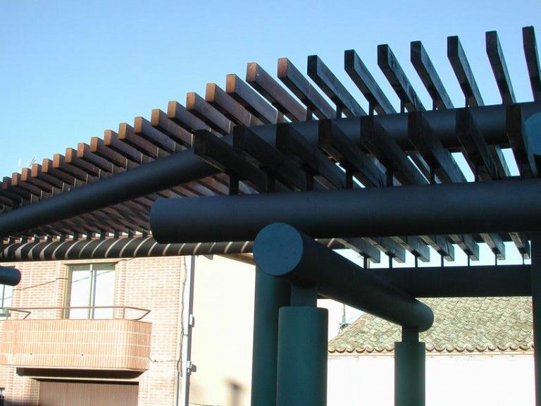 Pérgola con listones de madera oscura y soportes de color verde azulado contra un cielo azul.
