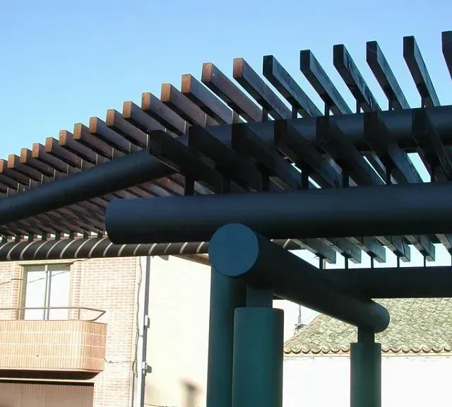 Estructura de pérgola de madera de color oscuro contra un cielo azul, parte de un edificio en el fondo.