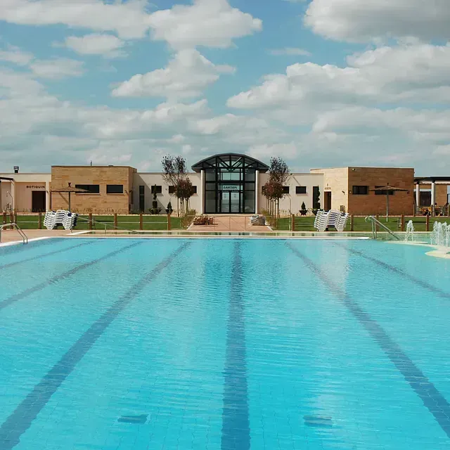 Una gran piscina al aire libre frente a un edificio moderno bajo un cielo azul con nubes blancas.
