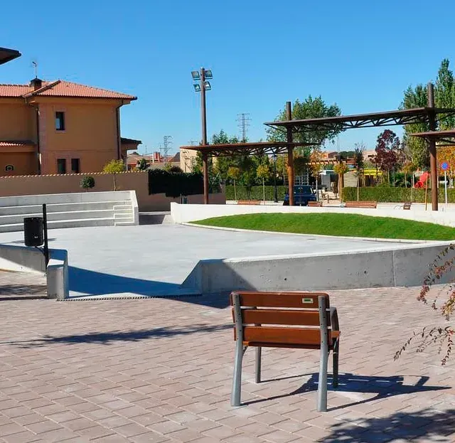 Un parque soleado con un escenario de hormigón, bancos y una zona de juegos frente a las casas.