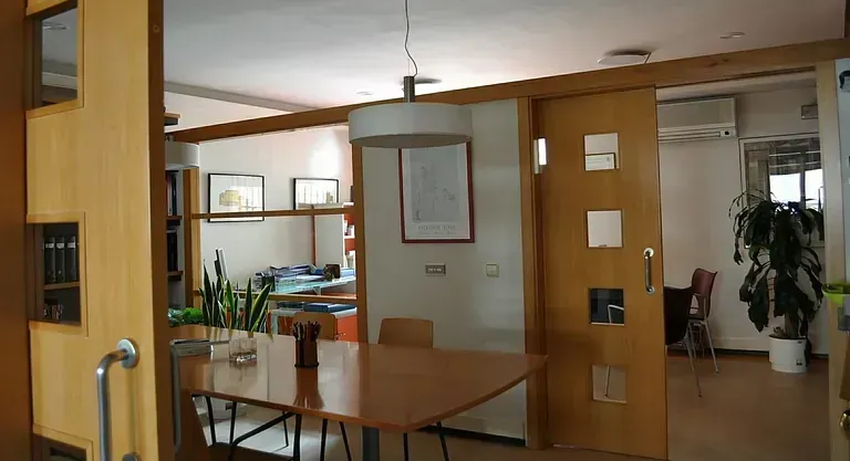 Oficina con paneles de madera, mesa, plantas y puerta que da a otra habitación con más plantas.