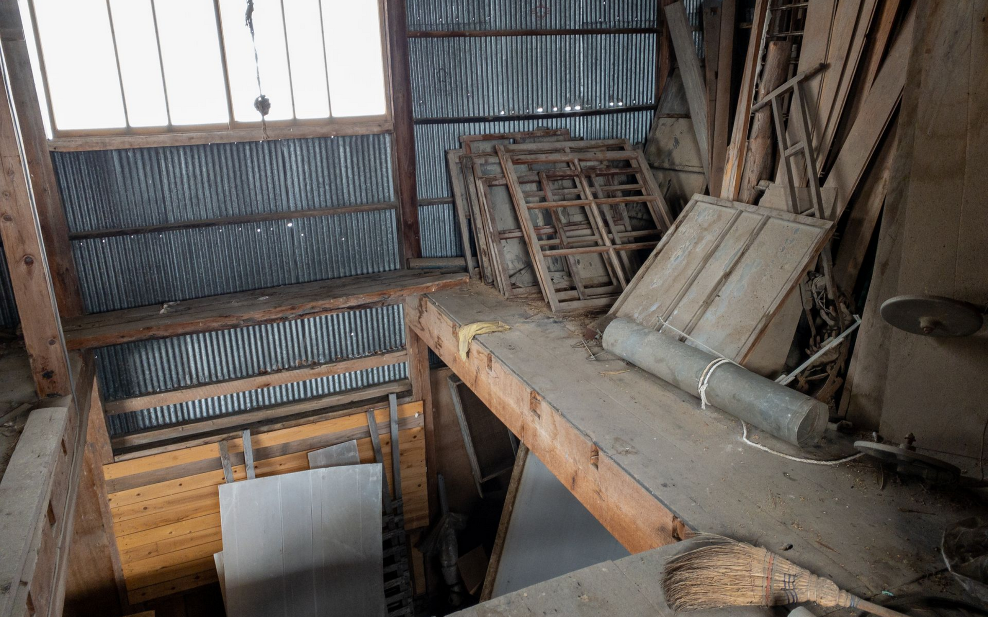 Vue intérieure d'un grenier encombré avec un toit en tôle ondulée, des poutres en bois et divers objets empilés contre les murs.