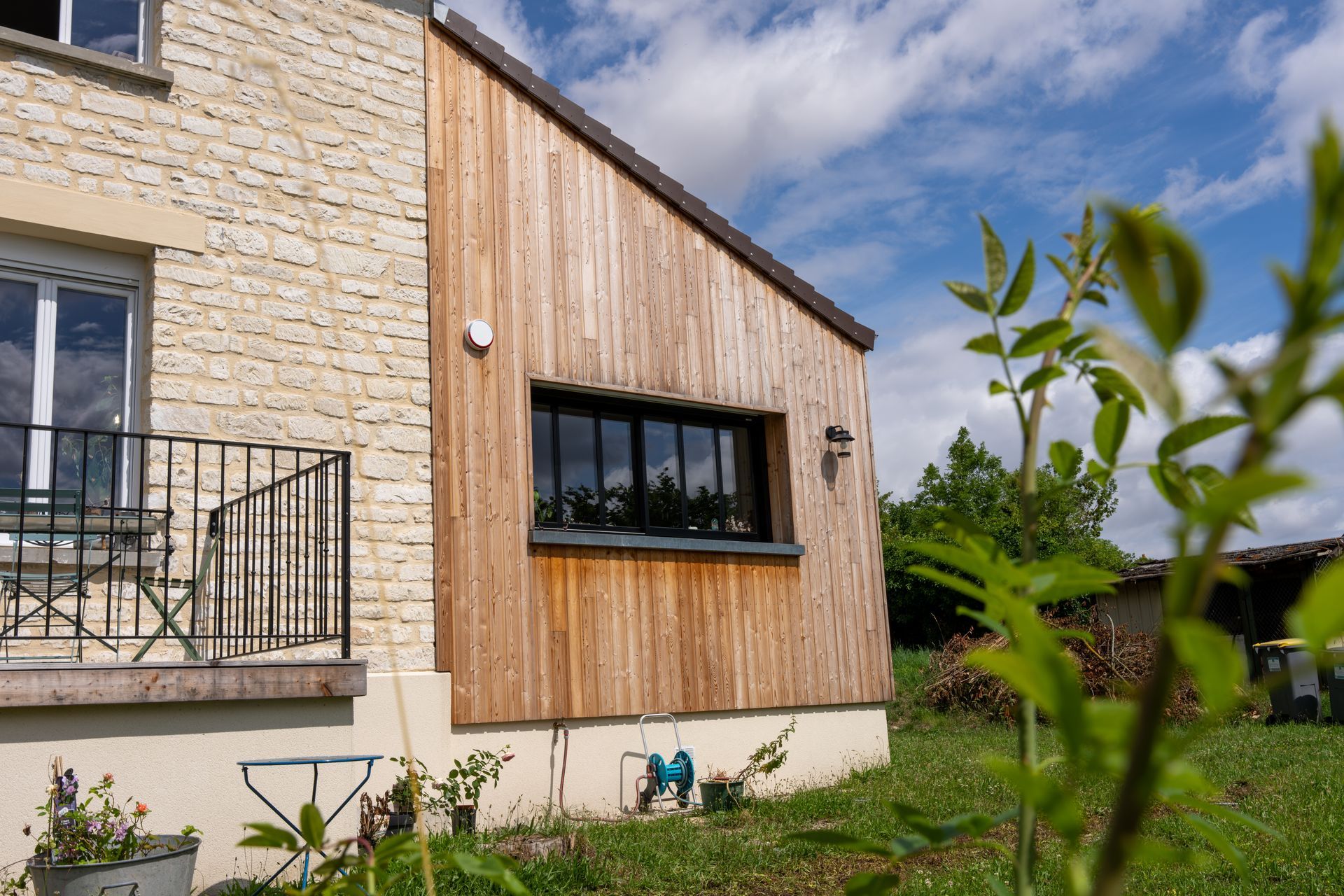 Une maison en pierre agrémentée d'une extension moderne en bois avec une fenêtre rectangulaire, entourée de pelouse et de plantes vertes.