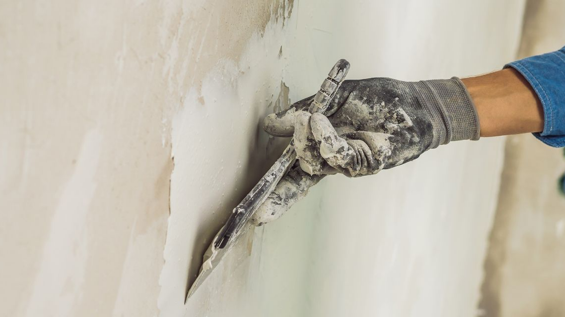 Une main gantée utilise un couteau à mastic métallique pour lisser du plâtre blanc sur un mur texturé.