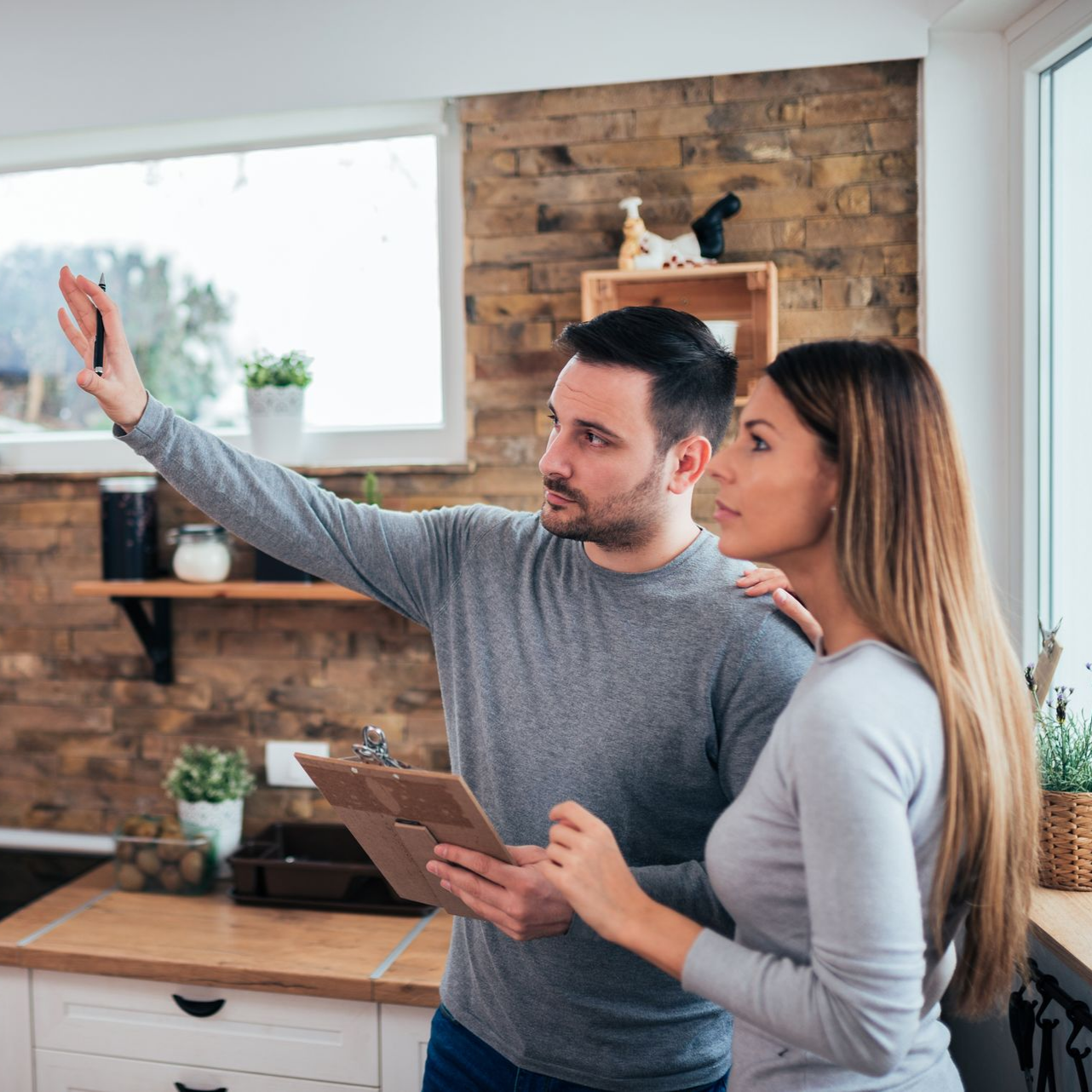 Un couple discute de travaux de rénovation dans une cuisine rustique ; la personne à gauche pointe du doigt en tenant un bloc-notes.