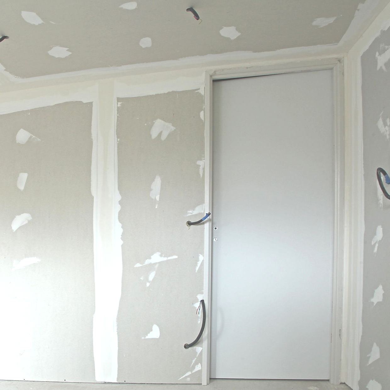Intérieur d'une pièce inachevée avec des panneaux de plaques de plâtre, des joints collés et une porte blanche installée dans un cadre métallique.