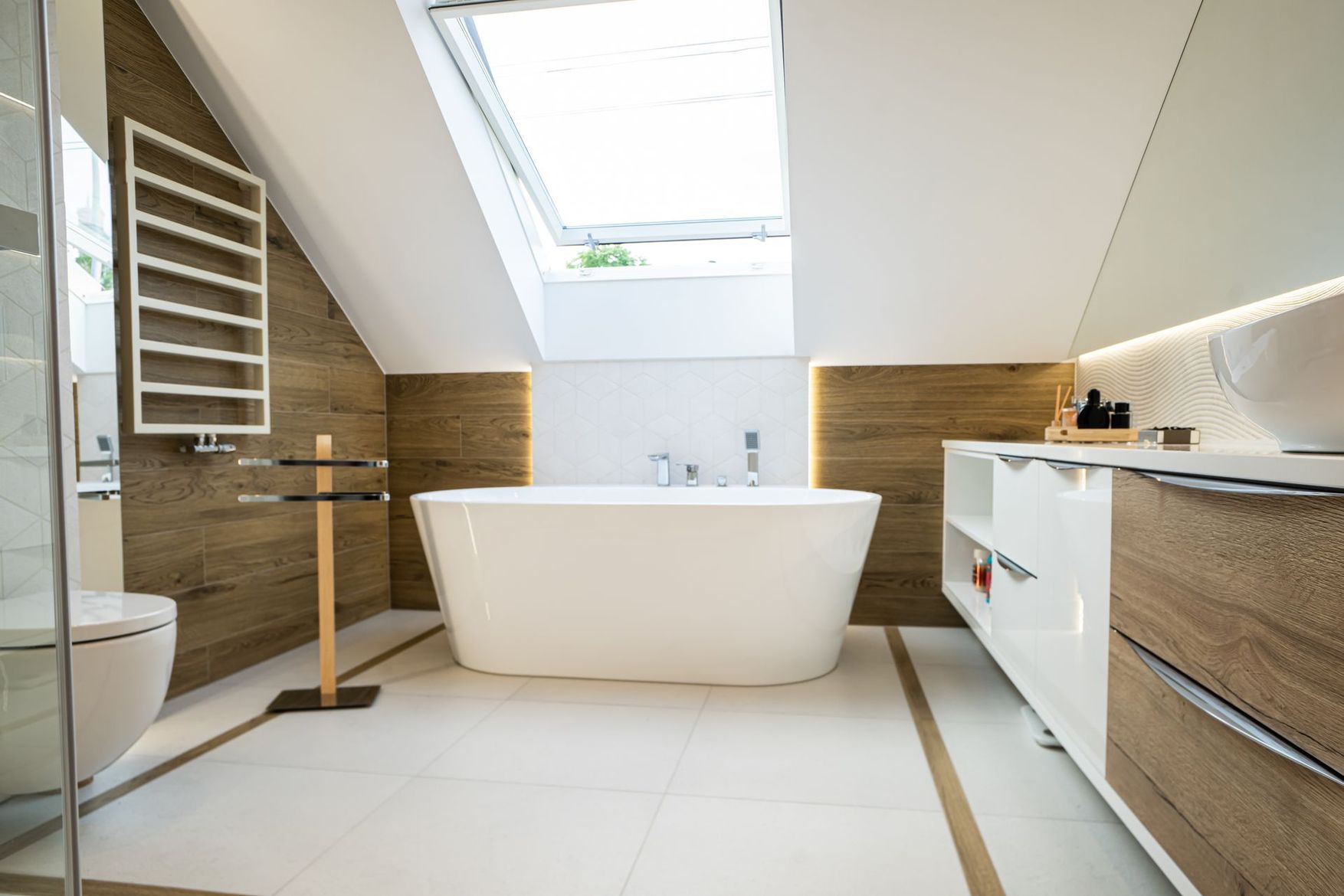 Salle de bains moderne avec baignoire blanche autoportante, murs lambrissés et puits de lumière sous un plafond en pente.