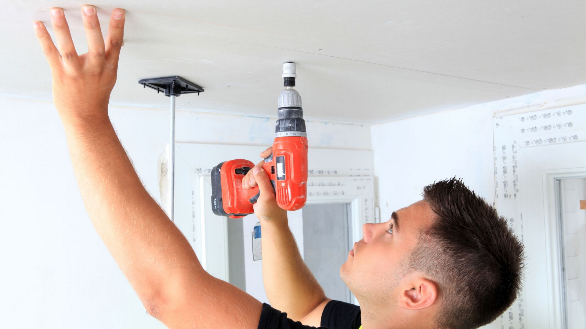 Une personne utilise une perceuse électrique rouge pour installer un panneau de plafond, en maintenant le panneau en place avec sa main gauche.