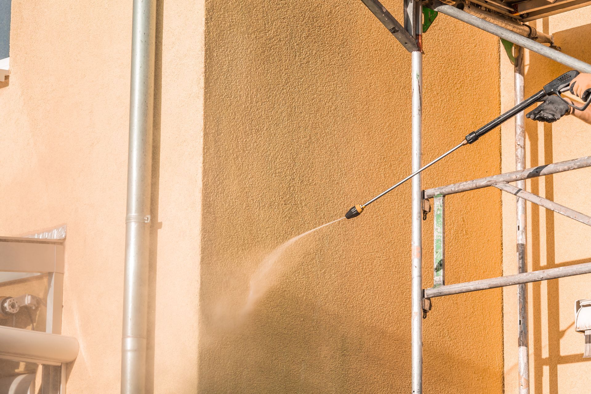 Un ouvrier sur un échafaudage utilise un nettoyeur à haute pression pour nettoyer un mur extérieur texturé de couleur beige.