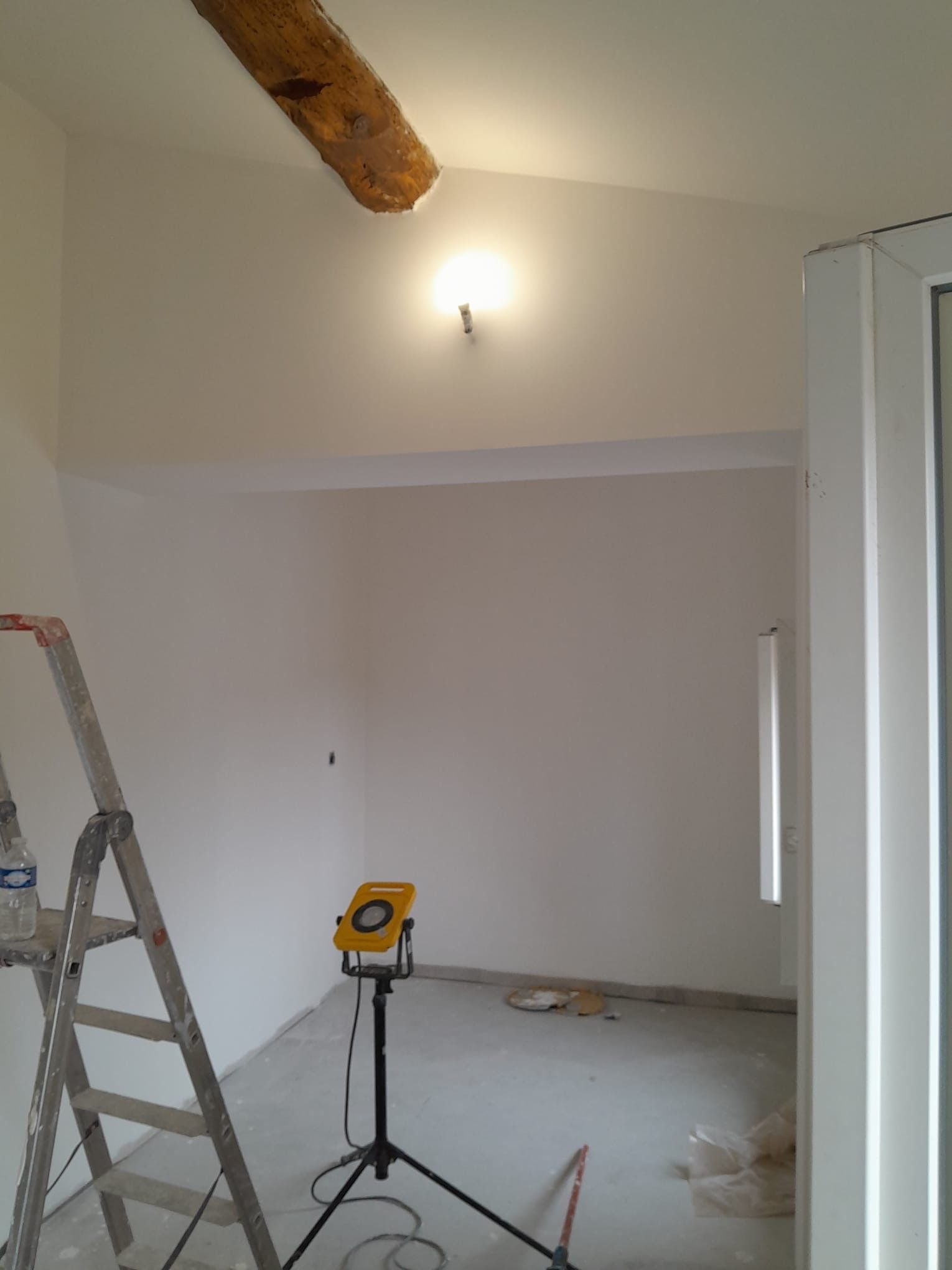 Une pièce inachevée aux murs blancs, avec une unique poutre de plafond en bois, une applique murale, un escabeau et une lampe de travail portative.
