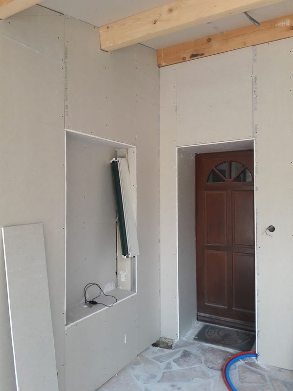 Pièce intérieure inachevée avec des cloisons sèches installées sur les murs, une niche murale rectangulaire et une porte en bois donnant sur l'extérieur.