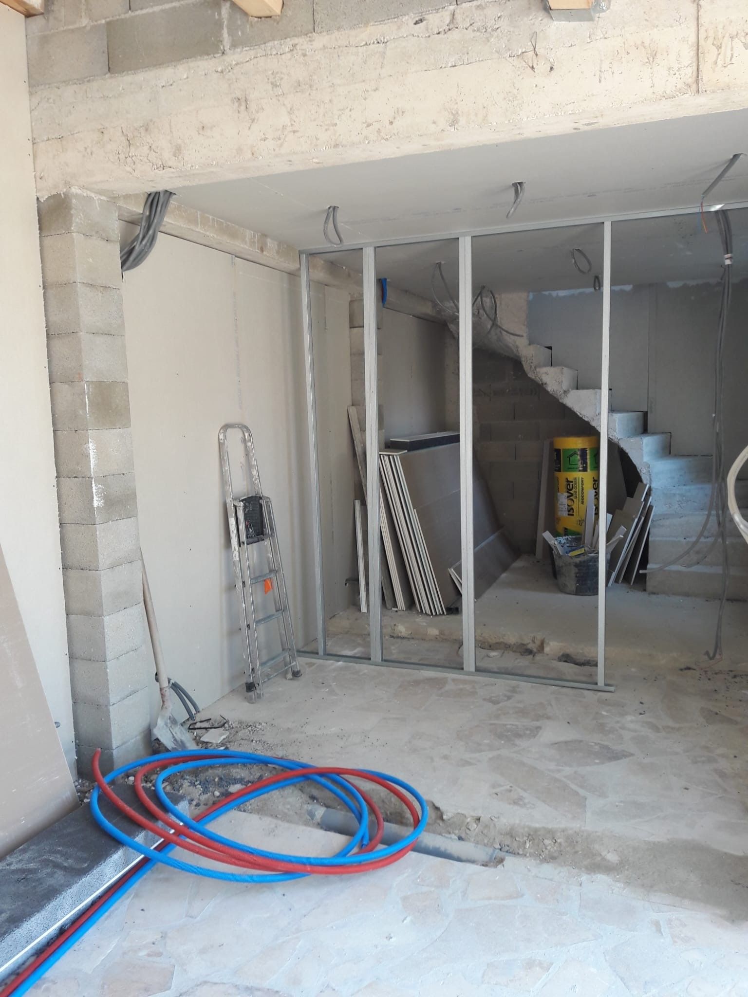 Un chantier de construction avec une ossature métallique, une échelle, des tuyaux bleus au sol et un escalier en arrière-plan.