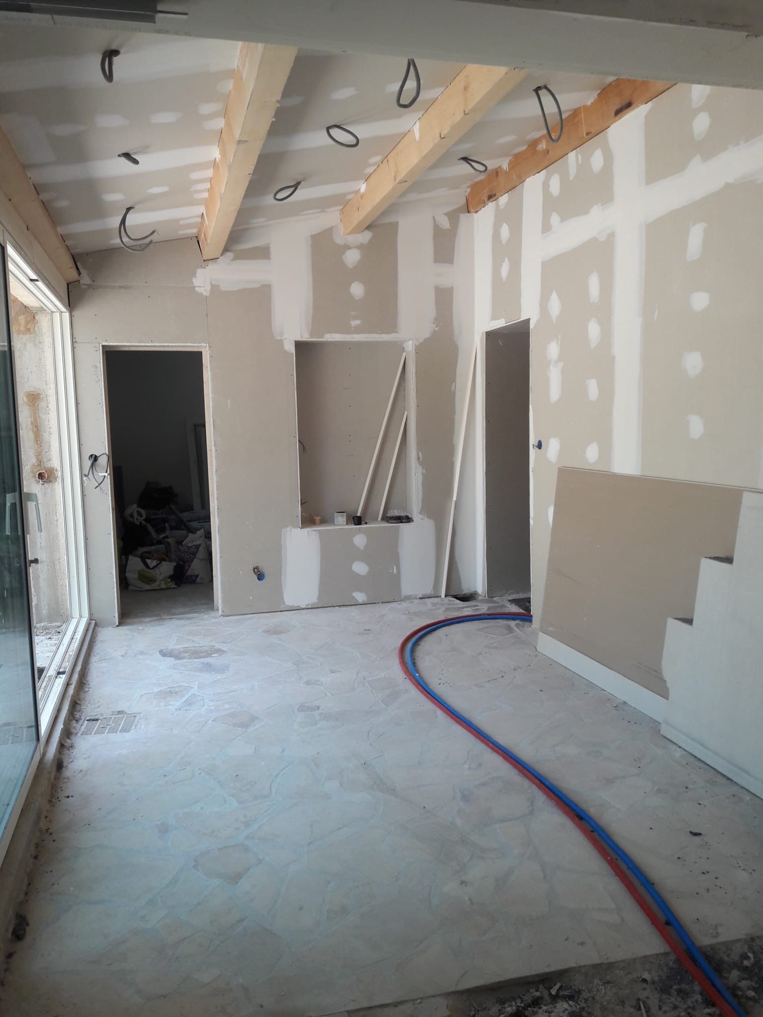 Une scène de construction intérieure montrant des murs en plaques de plâtre non peintes, des poutres de plafond en bois apparentes et un sol carrelé en pierre.