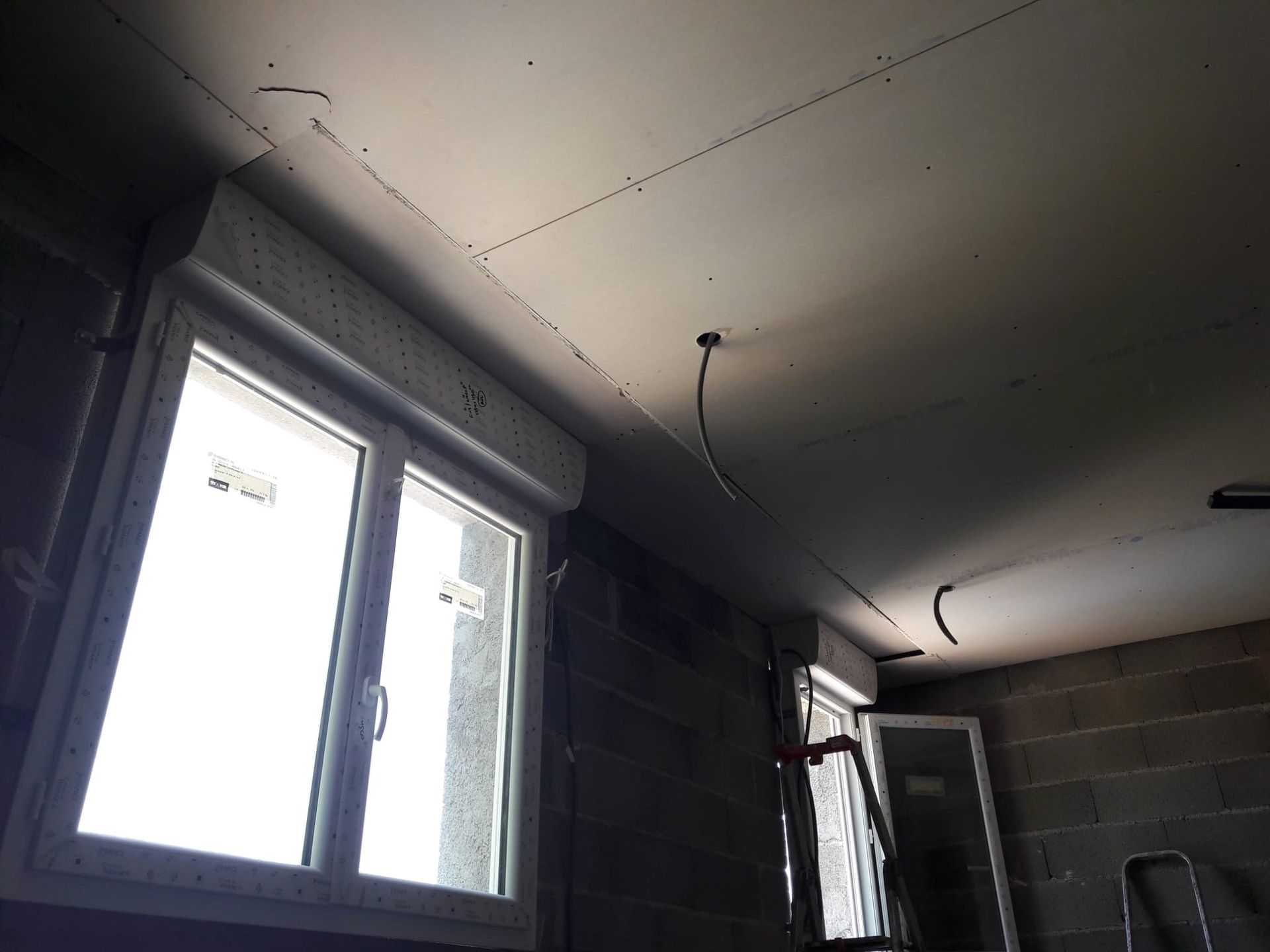 Intérieur d'une pièce inachevée présentant un plafond avec des joints de plaques de plâtre apparents et des fils électriques pendant au-dessus de deux fenêtres.