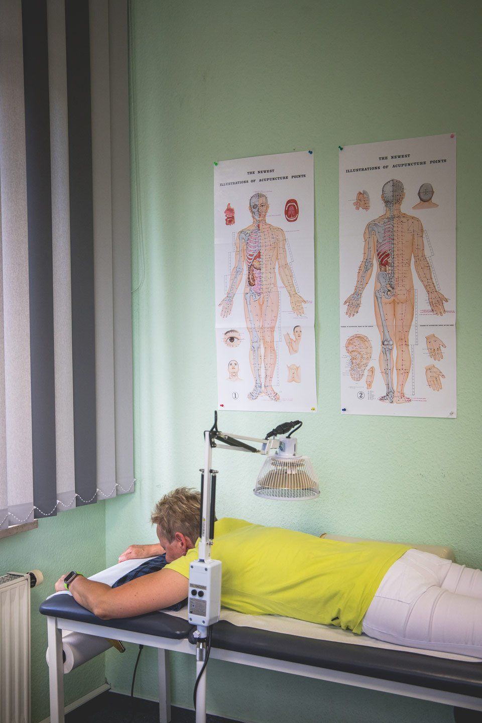 Person liegt auf einem Therapietisch unter einer Lampe in einem hellgrünen Raum mit anatomischen Postern.