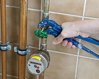 Apretando a mano una válvula azul en un contador de gas junto a tuberías de cobre y una pared alicatada.