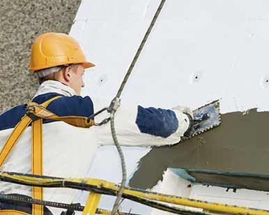 Un trabajador con casco aplica una capa de pintura blanca a una pared con una llana desde un andamio.