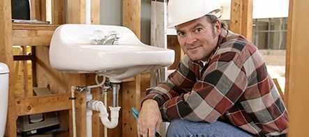 Un fontanero arrodillado junto a un lavabo blanco en un baño en construcción, con casco y camisa a cuadros.