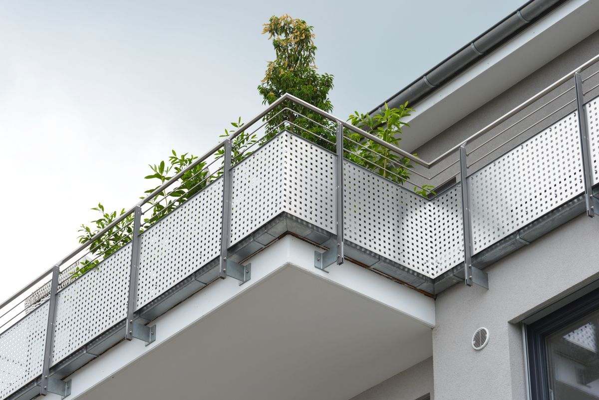 Balkon mit perforiertem Metallgeländer, silbernen Stützen, Pflanzen und hellgrauem Gebäude.