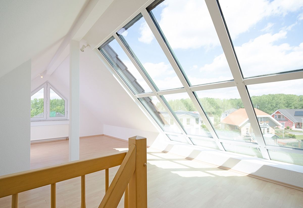 Innenraum eines hellen Dachzimmers mit großem Schrägfenster, Holzboden und Geländer sowie Blick auf Häuser und Bäume.