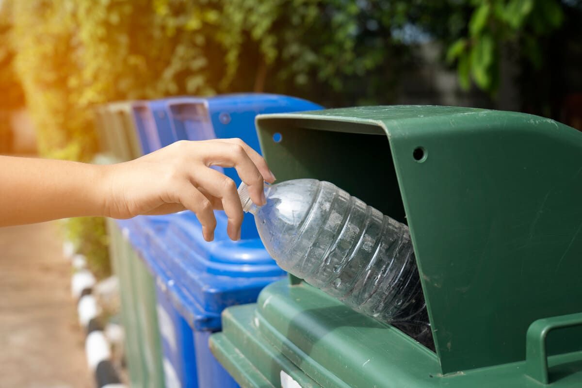 Mano colocando una botella de agua de plástico en un contenedor de reciclaje verde al lado de uno azul, al aire libre.