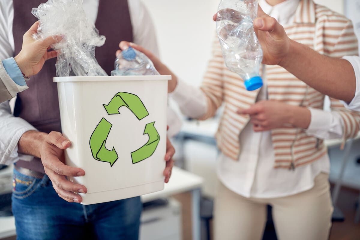 Personas que colocan desechos plásticos en un contenedor de reciclaje con un símbolo de reciclaje verde en un entorno de oficina.
